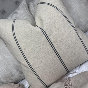 May include: A beige pillow with two dark gray stripes running diagonally across the front. The pillow has a textured fabric and is part of a set of pillows on a bed. The pillow is square-shaped.