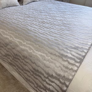 May include: A bed with a light gray textured bedspread featuring a wave-like pattern. Two decorative pillows with the word "Mrs." embroidered on them are visible. The bed is made up with a white bed sheet.
