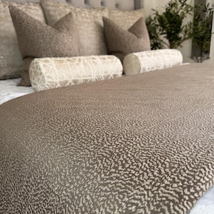 Puede incluir: Una cama con cabecero acolchado y varias almohadas decorativas. La colcha es de tela marrón texturizada con un patrón moteado. Dos almohadas cilíndricas con un diseño geométrico están frente a las almohadas.