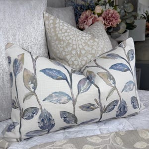 Puede incluir: Un cojín decorativo rectangular con un estampado de hojas botánicas en tonos azules, grises y beige. El cojín está sobre una colcha blanca acolchada, con otros cojines en el fondo.