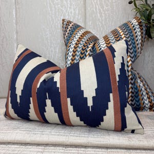 May include: A rectangular throw cushion featuring a bold navy blue and cream geometric pattern with vertical rust-coloured stripes, positioned in front of a second square throw cushion with a multi-coloured chevron pattern in blue, orange, and white.