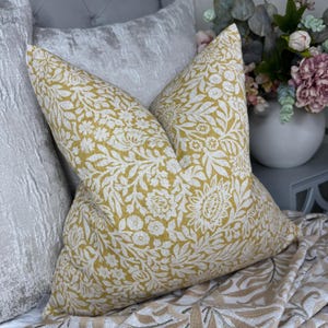 May include: Two decorative throw pillows with a floral pattern in yellow and white. The pillows are placed on a bed with a gray velvet headboard and a patterned blanket. The pillows have a soft, inviting appearance.