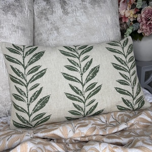 May include: Rectangular decorative pillow with a cream-colored background and a repeating pattern of dark green leaf branches. The pillow is placed on a bed with a patterned beige and white blanket.