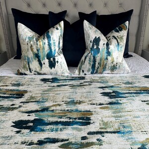 May include: A bed with a tufted headboard, featuring navy blue and patterned throw pillows. The patterned pillows have a white background with blue, teal, and gold brushstroke designs. A matching bed runner with similar brushstroke patterns completes the ensemble.