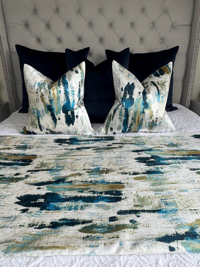 May include: A bed with a tufted headboard, featuring navy blue and patterned throw pillows. The patterned pillows have a white background with blue, teal, and gold brushstroke designs. A matching bed runner with similar brushstroke patterns completes the ensemble.