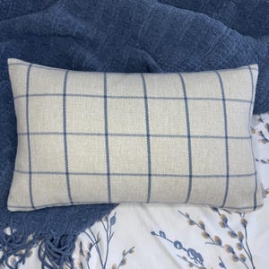 May include: Rectangular pillow with a cream-colored background and a blue plaid pattern. The pillow is placed on a blue blanket and a white floral patterned sheet. The pillow's design features a grid of intersecting blue lines.