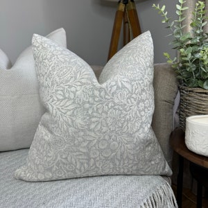 May include: A white and grey patterned cushion with a floral design. The cushion is on a grey sofa with a fringed throw blanket.