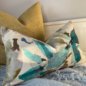 May include: Two decorative throw pillows, one with a mustard yellow fabric and the other with a white background and abstract blue, green, and brown watercolor-like patterns.