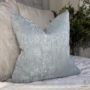 Può includere: Un cuscino blu chiaro con una superficie testurizzata e screpolata. Il cuscino è realizzato in un tessuto morbido e lussuoso e ha una lucentezza discreta.
