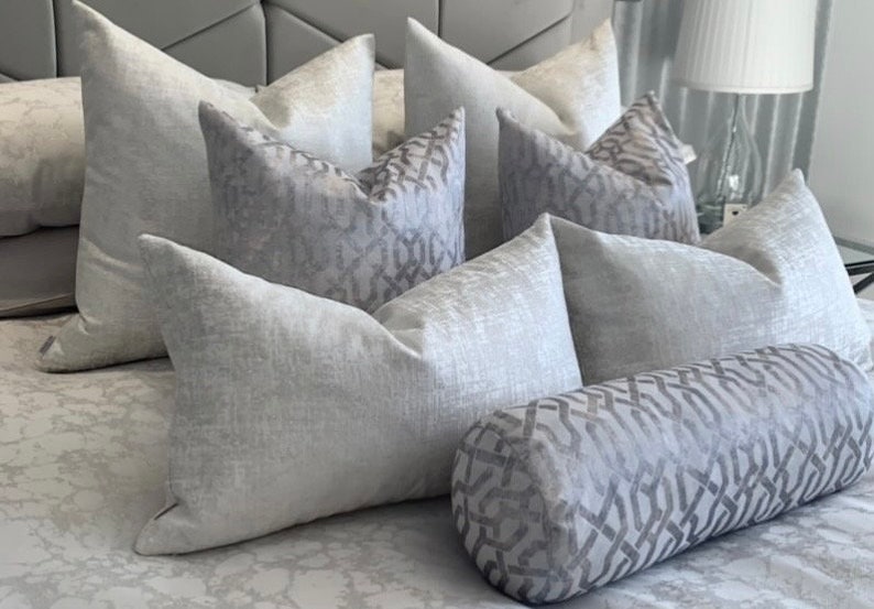 Pu&ograve; includere: Una collezione di cuscini decorativi grigi e bianchi con un motivo con un cuscino di supporto grigio e bianco con un motivo. I cuscini sono disposti su un copriletto grigio e bianco con un motivo.