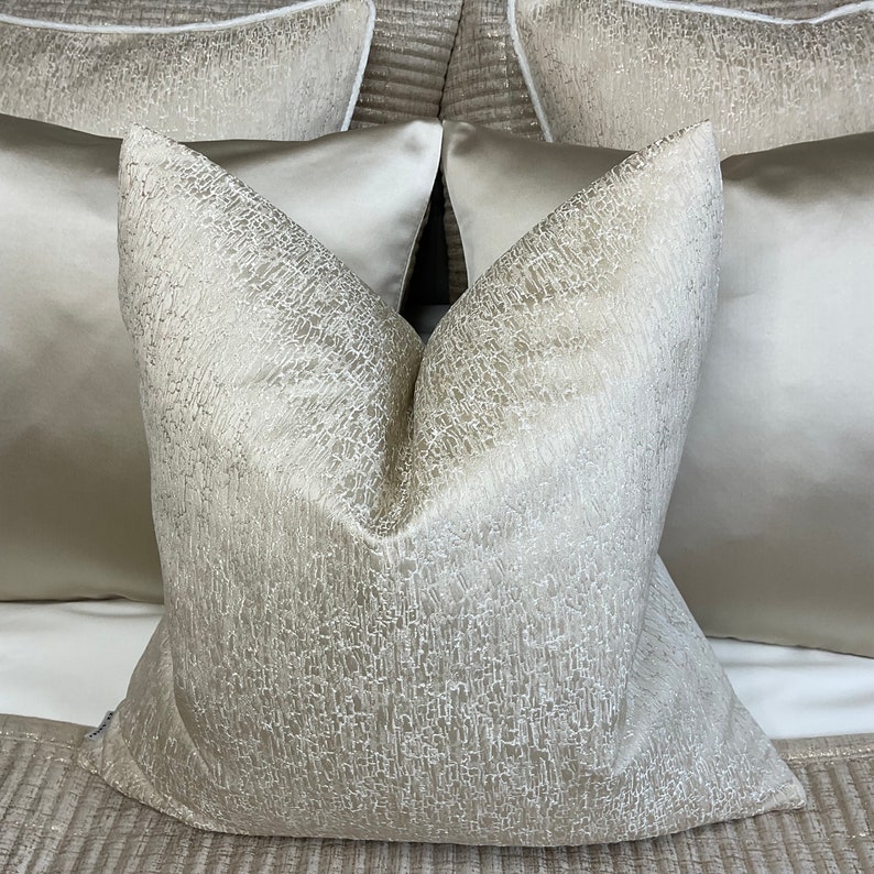 May include: Two decorative throw pillows with a textured, shimmery fabric in a light beige color. The pillows are arranged on a bed with a textured, beige bedspread.