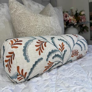May include: A cylindrical decorative pillow with a cream-colored linen fabric. The pillow features an embroidered design of blue and rust-colored leaves and vines. The pillow is placed on a white bedspread with other decorative pillows in the background.