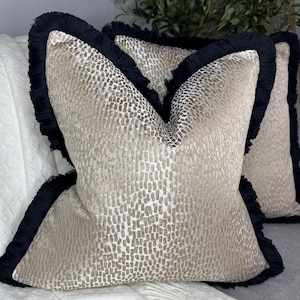 K&ouml;nnte beinhalten: Zwei dekorative Kissen mit einem geometrischen Muster in Beige und Gold. Die Kissen haben einen schwarzen Fransenbesatz an den R&auml;ndern.
