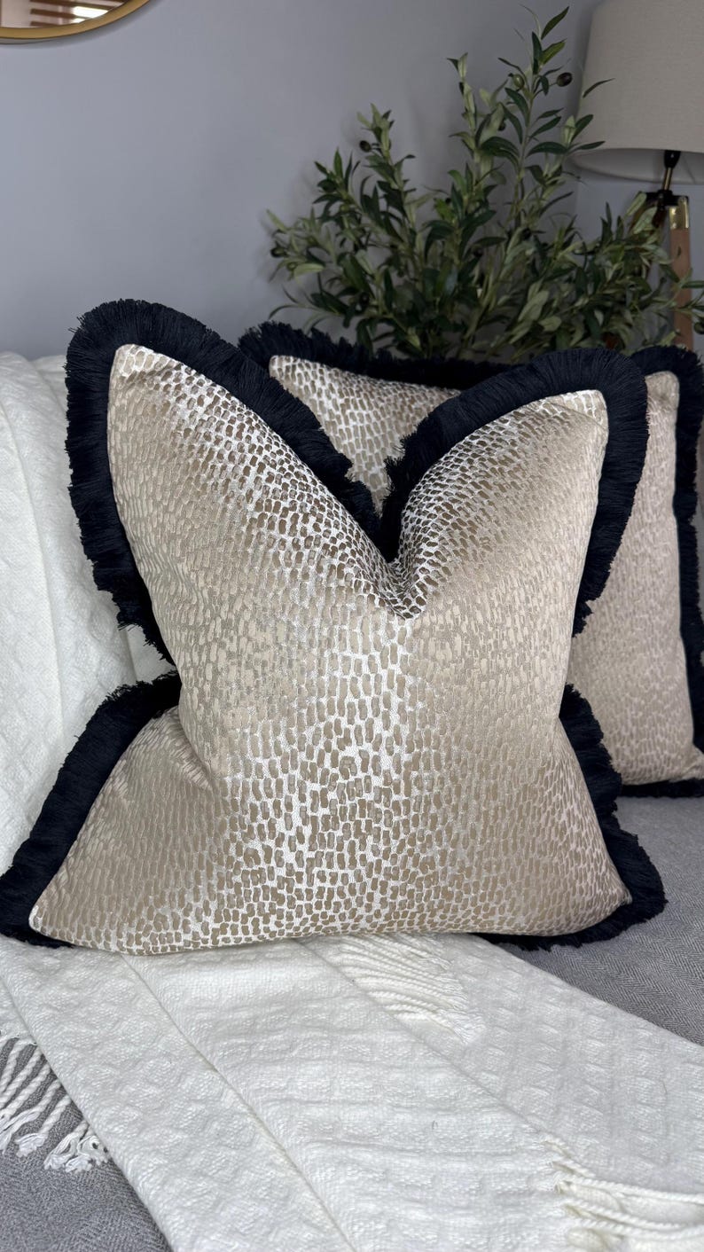 K&ouml;nnte beinhalten: Zwei dekorative Kissen mit einem geometrischen Muster in Beige und Gold. Die Kissen haben einen schwarzen Fransenbesatz an den R&auml;ndern.