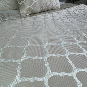 May include: A close-up of a beige bedspread with a white geometric pattern. The pattern is a repeating design of squares and circles.