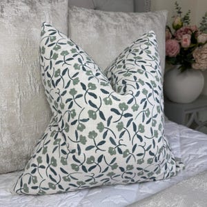 May include: A white throw pillow with a green and blue floral pattern. The pillow is made of a soft, linen-like fabric and has a subtle sheen.