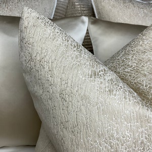 May include: Close-up of two decorative throw pillows with a textured, woven fabric. The pillows are a light beige color with a subtle shimmer.