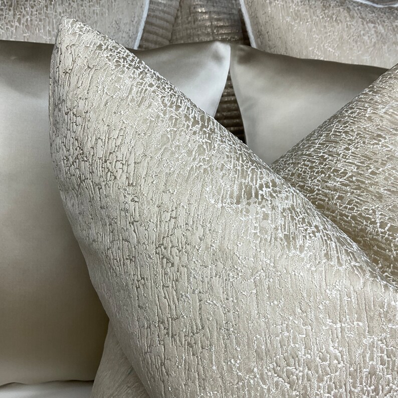 May include: Close-up of two decorative throw pillows with a textured, woven fabric. The pillows are a light beige color with a subtle shimmer.