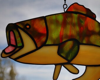 May include: A stained glass fish with orange, yellow, and green colors. The fish is hanging from a chain and has a black outline.
