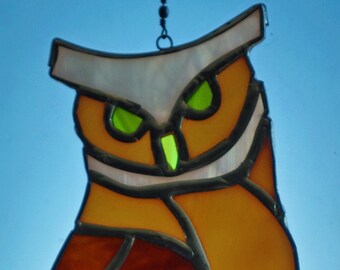 May include: A stained glass owl with brown, orange, and white glass pieces. The owl has green eyes and is hanging from a chain.
