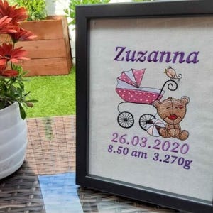 Op de afbeelding: Een ingelijste borduurwerk met de naam "Zuzanna" in paars, met een kinderwagen, teddybeer en vogel. De datum 26.03.2020, tijd 8.50 uur en gewicht 3,270g zijn geborduurd. De lijst is zwart.