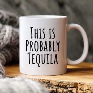 May include: White ceramic mug with the text "THIS IS PROBABLY TEQUILA" in black lettering. The mug has a curved handle and sits on a wooden surface. A gray knitted blanket is in the background.