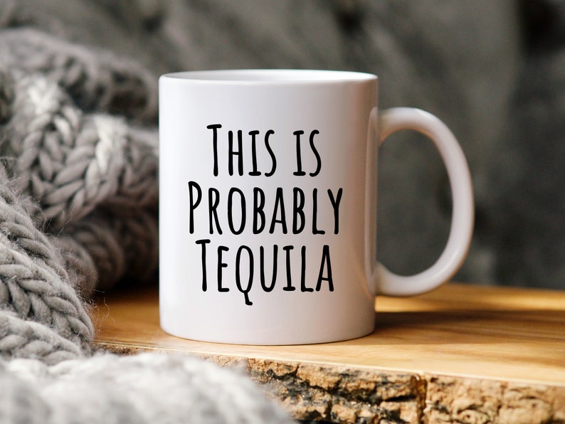 May include: White ceramic mug with the text "THIS IS PROBABLY TEQUILA" in black lettering. The mug has a curved handle and sits on a wooden surface. A gray knitted blanket is in the background.