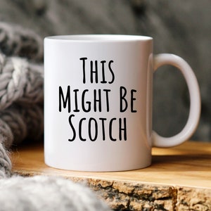 May include: White ceramic mug with the text "THIS MIGHT BE SCOTCH" in black. The mug has a curved handle and is set on a wooden surface. A gray knitted blanket is in the background.