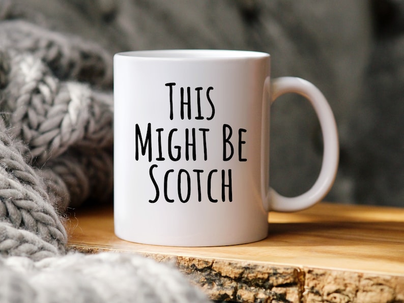 May include: White ceramic mug with the text "THIS MIGHT BE SCOTCH" in black. The mug has a curved handle and is set on a wooden surface. A gray knitted blanket is in the background.