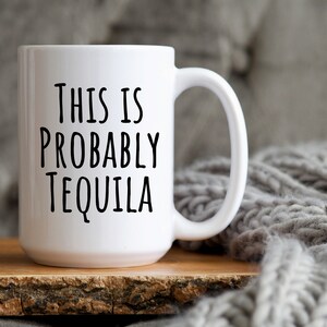 May include: White ceramic mug with the text "THIS IS PROBABLY TEQUILA" in black, bold letters. The mug has a large handle and sits on a wooden surface, with a gray knitted blanket in the background.