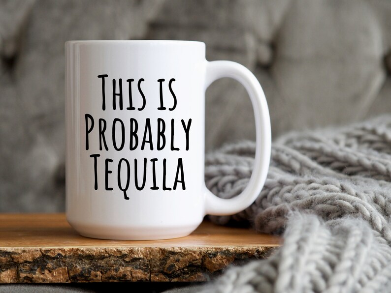 May include: White ceramic mug with the text "THIS IS PROBABLY TEQUILA" in black, bold letters. The mug has a large handle and sits on a wooden surface, with a gray knitted blanket in the background.