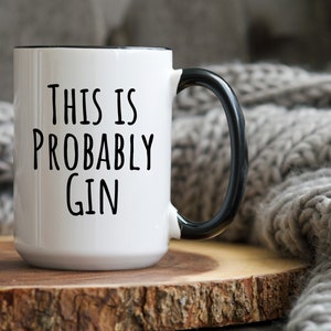 May include: White ceramic mug with a black handle and rim. The mug features the text "THIS IS PROBABLY GIN" in black, bold letters. The mug is set on a wooden surface.