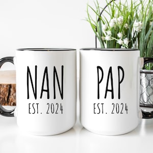 May include: Two white ceramic mugs with black rims and handles. The mug on the left says "NAN EST. 2024" and the mug on the right says "PAP EST. 2024".