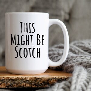 May include: A white ceramic mug with the text "THIS MIGHT BE SCOTCH" in black, sans-serif font. The mug sits on a wooden surface, with a gray knitted blanket in the background. The mug has a large handle.