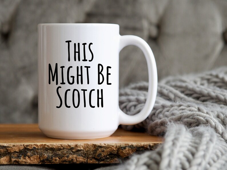 May include: A white ceramic mug with the text "THIS MIGHT BE SCOTCH" in black, sans-serif font. The mug sits on a wooden surface, with a gray knitted blanket in the background. The mug has a large handle.