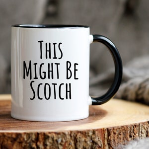 May include: White ceramic mug with a black interior and handle. The mug features the text "THIS MIGHT BE SCOTCH" in black, bold letters. The mug is set on a wooden surface, with a soft, gray textile in the background.
