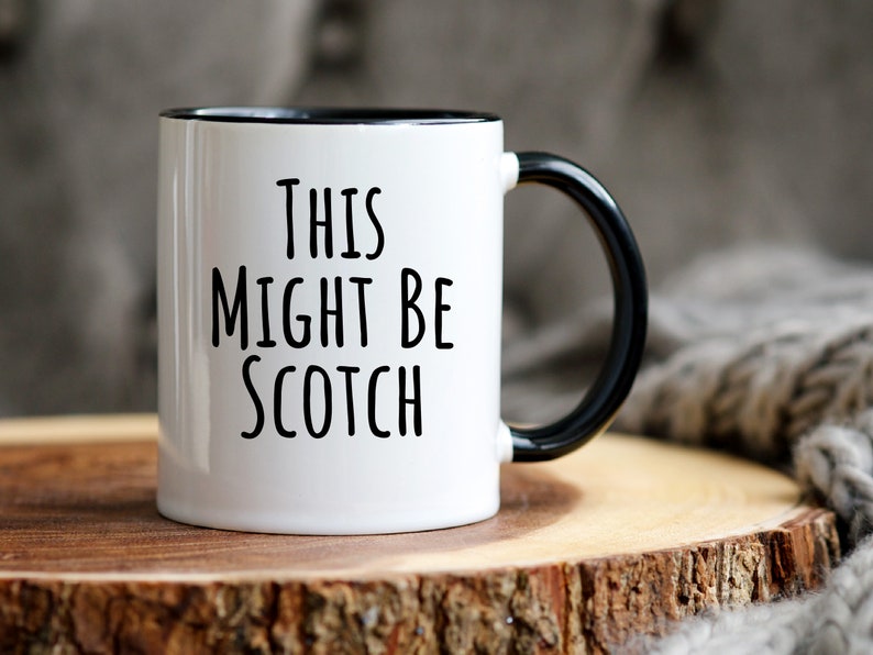 May include: White ceramic mug with a black interior and handle. The mug features the text "THIS MIGHT BE SCOTCH" in black, bold letters. The mug is set on a wooden surface, with a soft, gray textile in the background.
