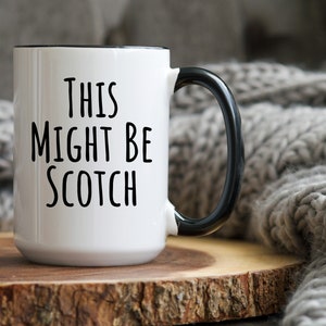 May include: White ceramic mug with black handle and rim. The mug features the text "THIS MIGHT BE SCOTCH" in black, bold letters. The mug is placed on a wooden surface.