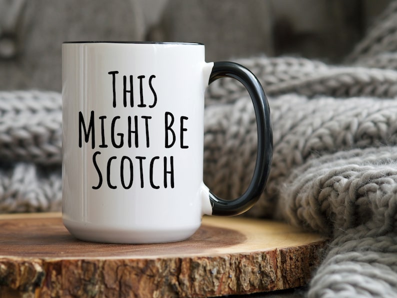 May include: White ceramic mug with black handle and rim. The mug features the text "THIS MIGHT BE SCOTCH" in black, bold letters. The mug is placed on a wooden surface.