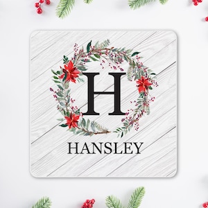 May include: A square coaster with a white wood grain background features a floral wreath with red poinsettias and greenery around a large black letter "H". The name "HANSLEY" is printed in black below the letter.