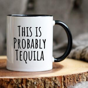 May include: White ceramic mug with a black interior and handle. The mug features the text "THIS IS PROBABLY TEQUILA" in black, bold letters. The mug is set on a wooden surface.