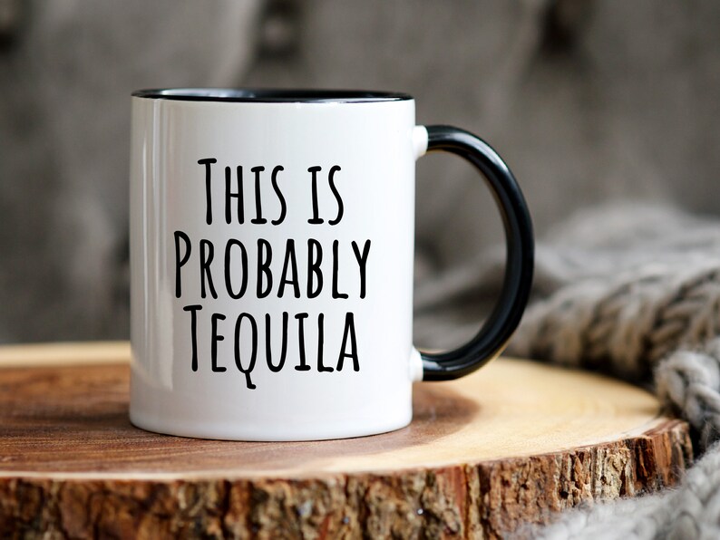 May include: White ceramic mug with a black interior and handle. The mug features the text "THIS IS PROBABLY TEQUILA" in black, bold letters. The mug is set on a wooden surface.