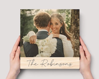May include: A square wooden plaque featuring a printed photograph of a couple embracing, with the person on the right holding a bouquet of white flowers. The bottom section of the plaque displays the text The Robinsons in black script font.
