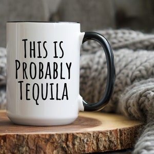 May include: White ceramic mug with a black handle and rim. The mug features the text "THIS IS PROBABLY TEQUILA" in black, bold letters. The mug is set on a wooden surface against a blurred gray background.