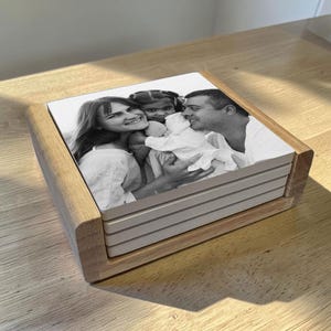 May include: A stack of four square ceramic coasters held in a light-toned wooden holder. The top coaster features a black and white photograph of a smiling woman, a man, and a young child embracing, with the image printed across the surface.