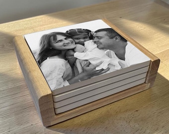May include: A stack of four square ceramic coasters held in a light-toned wooden holder. The top coaster features a black and white photograph of a smiling woman, a man, and a young child embracing, with the image printed across the surface.