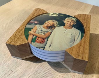 May include: A stack of circular coasters featuring a photograph of two children, held within a light brown wooden holder. The top coaster displays a girl in a grey shirt and a boy in a long-sleeved white shirt. The holder has a geometric design.