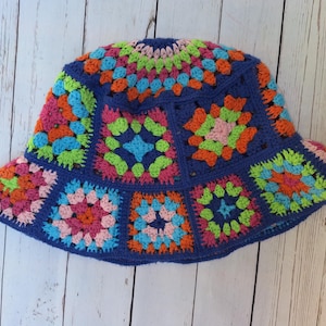 May include: A crochet bucket hat constructed from multiple colorful granny squares joined by a dark blue yarn border. The hat features a vibrant pattern of pink, orange, light blue, and lime green yarn, creating a geometric design throughout the crown.