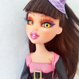 Könnte beinhalten: Eine Puppe mit braunen Haaren, die ein rosa Kleid mit einem schwarzen Gürtel und einen lila Hexenhut trägt. Die Puppe hat gelbe Augen und orangefarbenes Lidschatten.