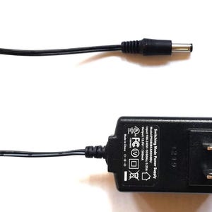 May include: A black switching mode power supply with a two-pin plug and a DC connector. The power supply has text with the model number, input, and output specifications. The DC connector is connected to a black cable.